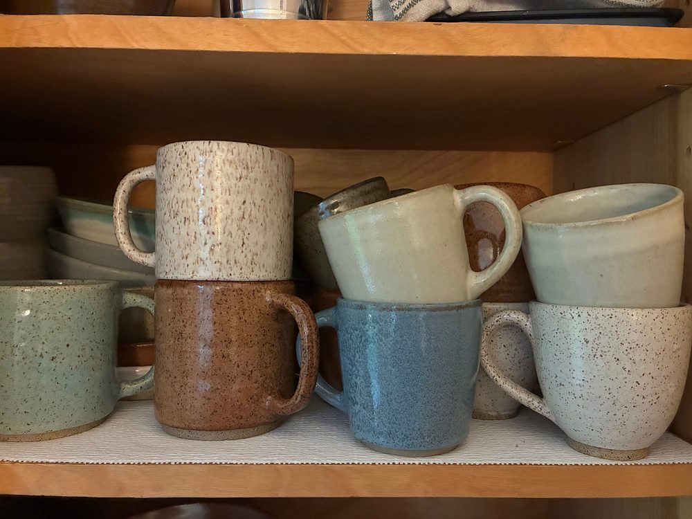 A selection of cups in speckled white, brown, green and blue glazes, stacked on a kitchen shelf