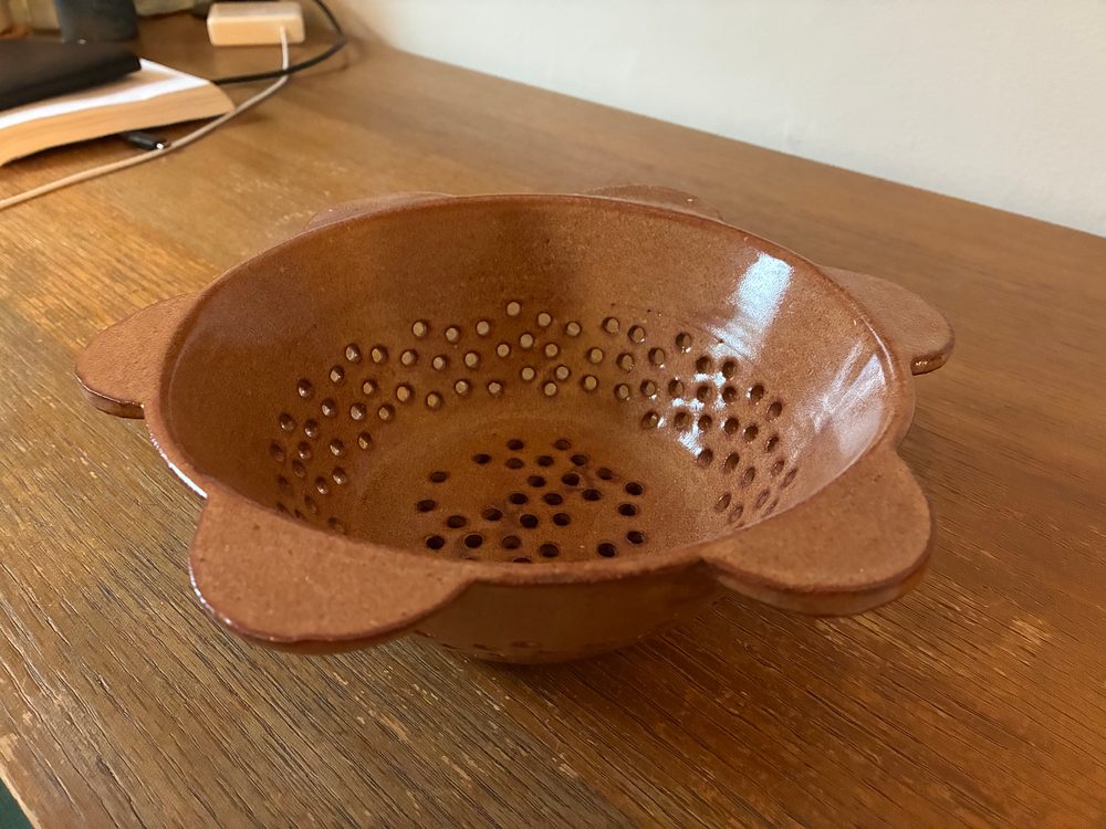 The daisy colander, nutmeg glaze on brown clay, with petal-shaped handles around the rim and holes in a flower pattern